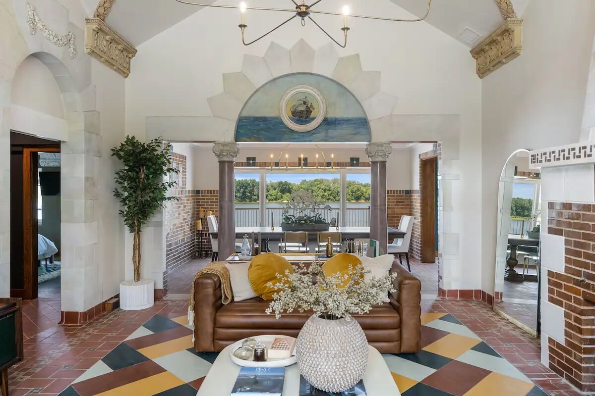 Spacious living room with a leather sofa and decorative pillows. A dining area is visible in the background through an archway. The room features patterned tiles, a potted plant, and a vaulted ceiling.