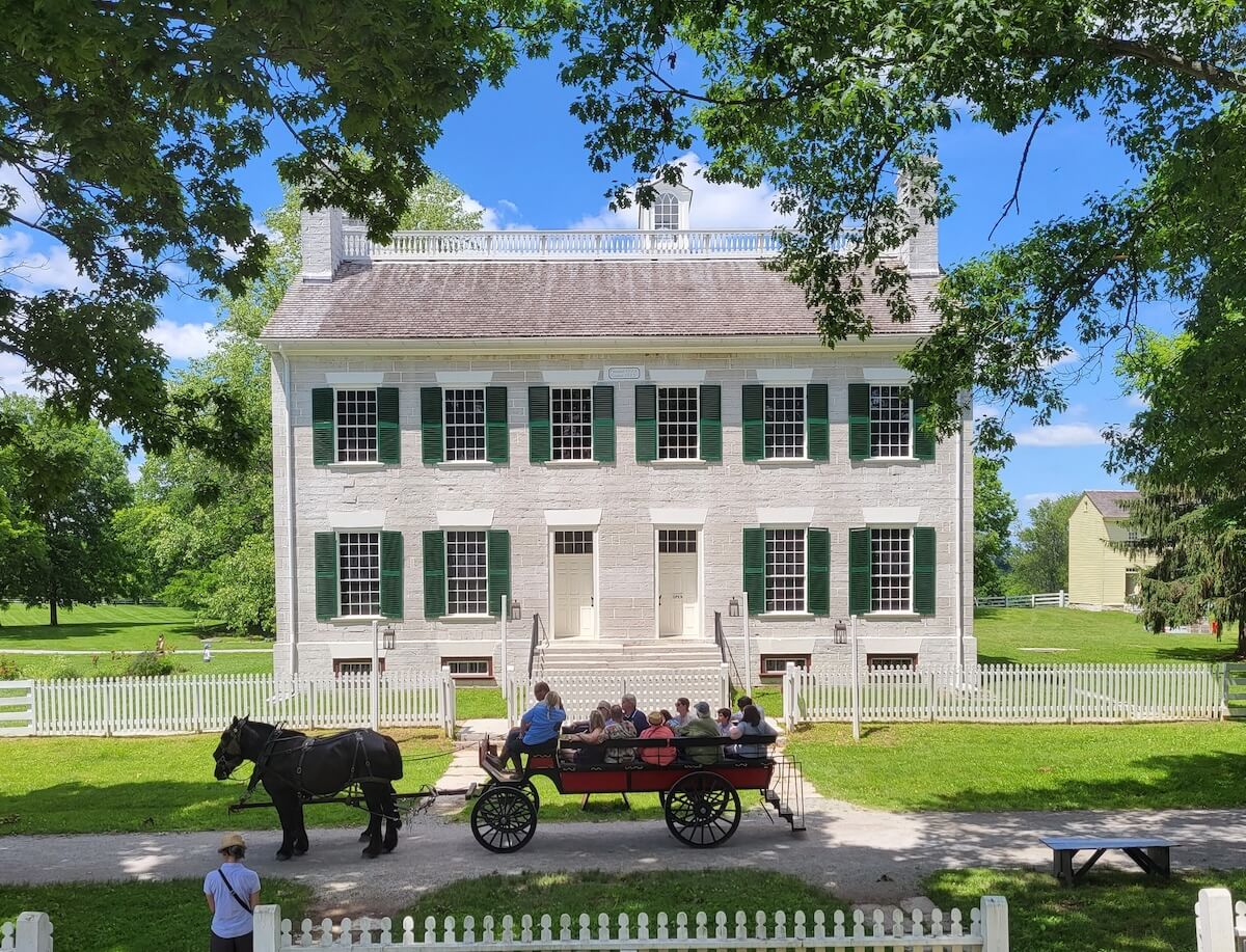A horse-drawn carriage with passengers is parked in front of a large two-story white building with green shutters on its windows. The charming scene, set in a leafy area with a white picket fence, offers one of those unforgettable KY stays that should be on everyone's bucket list.