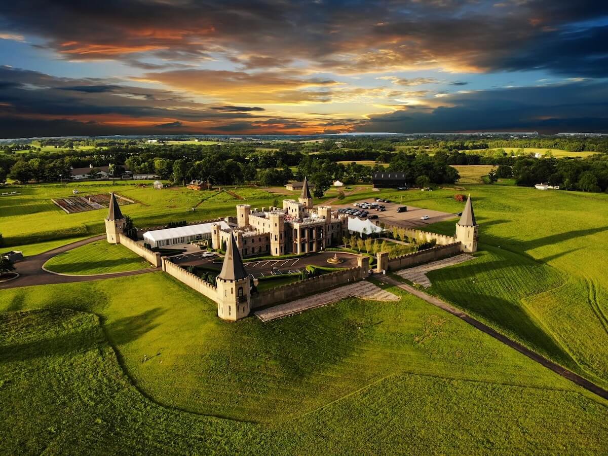 An aerial view of a large, castle-like mansion with four corner towers, surrounded by green fields and a dramatic sky at sunset.