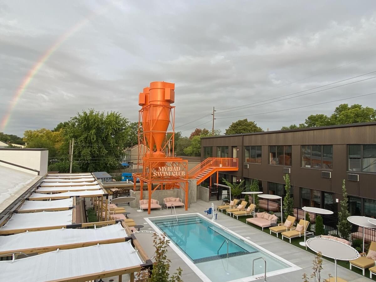 A swimming pool and charming lounge area sit next to a modern building. An orange, silo-shaped structure labeled "St. Miriam Swim Club" is prominent. A rainbow is faintly visible in the cloudy sky.