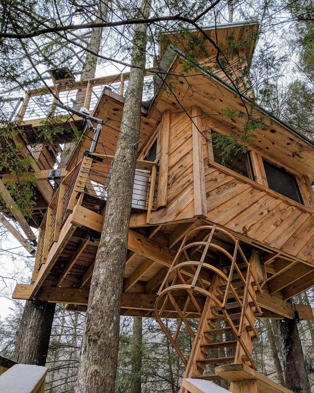 A multi-level wooden treehouse with a spiral staircase, built around several tall trees in a serene forest setting.