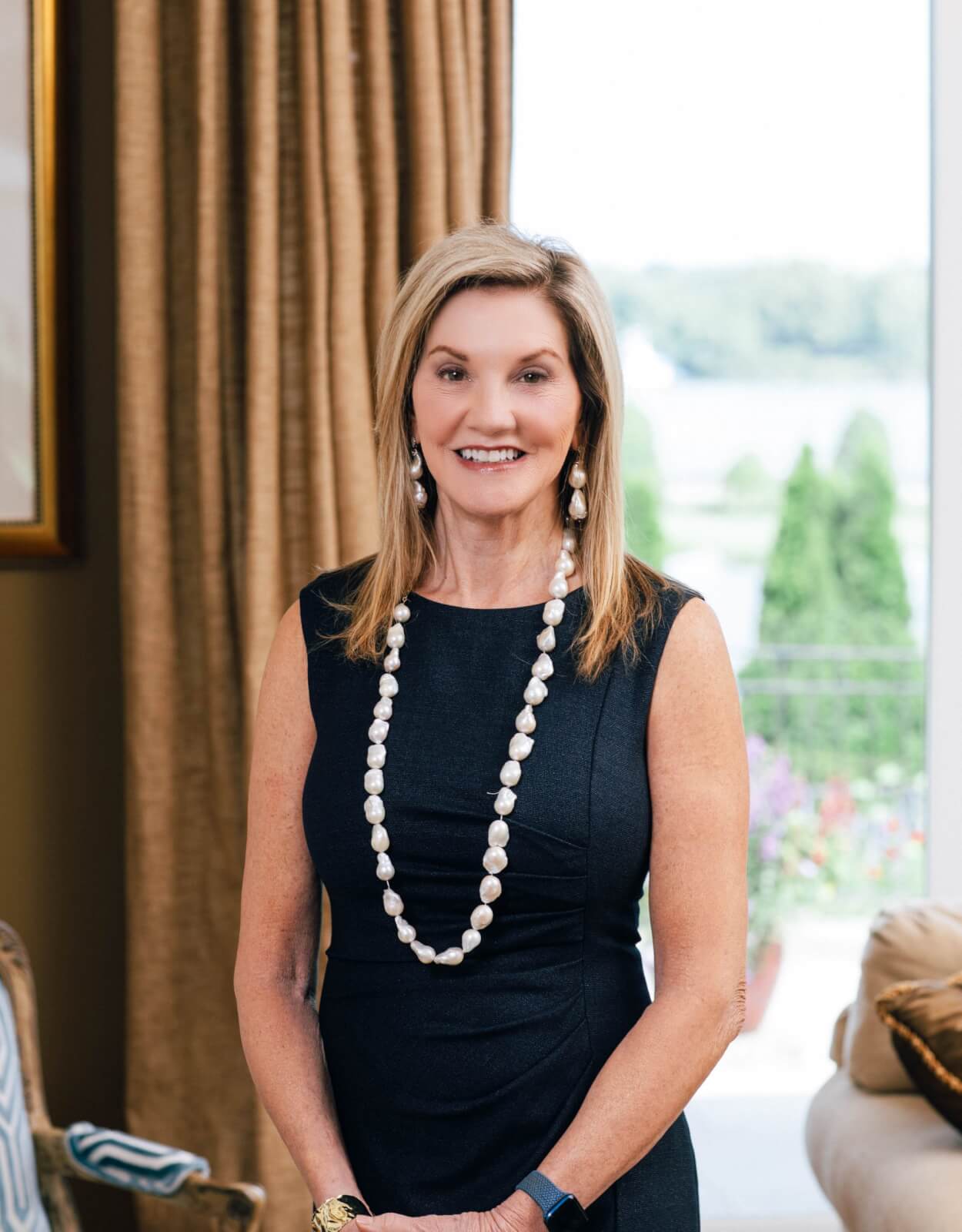 A woman in a sleeveless black dress, wearing a long beaded necklace and earrings, stands indoors with a relaxed smile. Beige curtains and a window with an outdoor view are in the background.