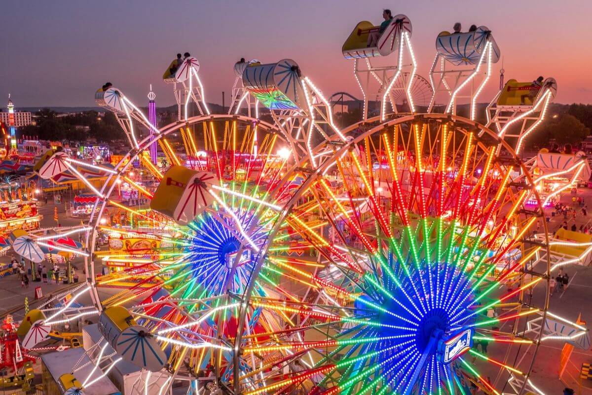A brightly lit carnival at dusk features two large Ferris wheels and numerous colorful lights, capturing the lively atmosphere of Louisville events in August, with people and attractions visible in the background.