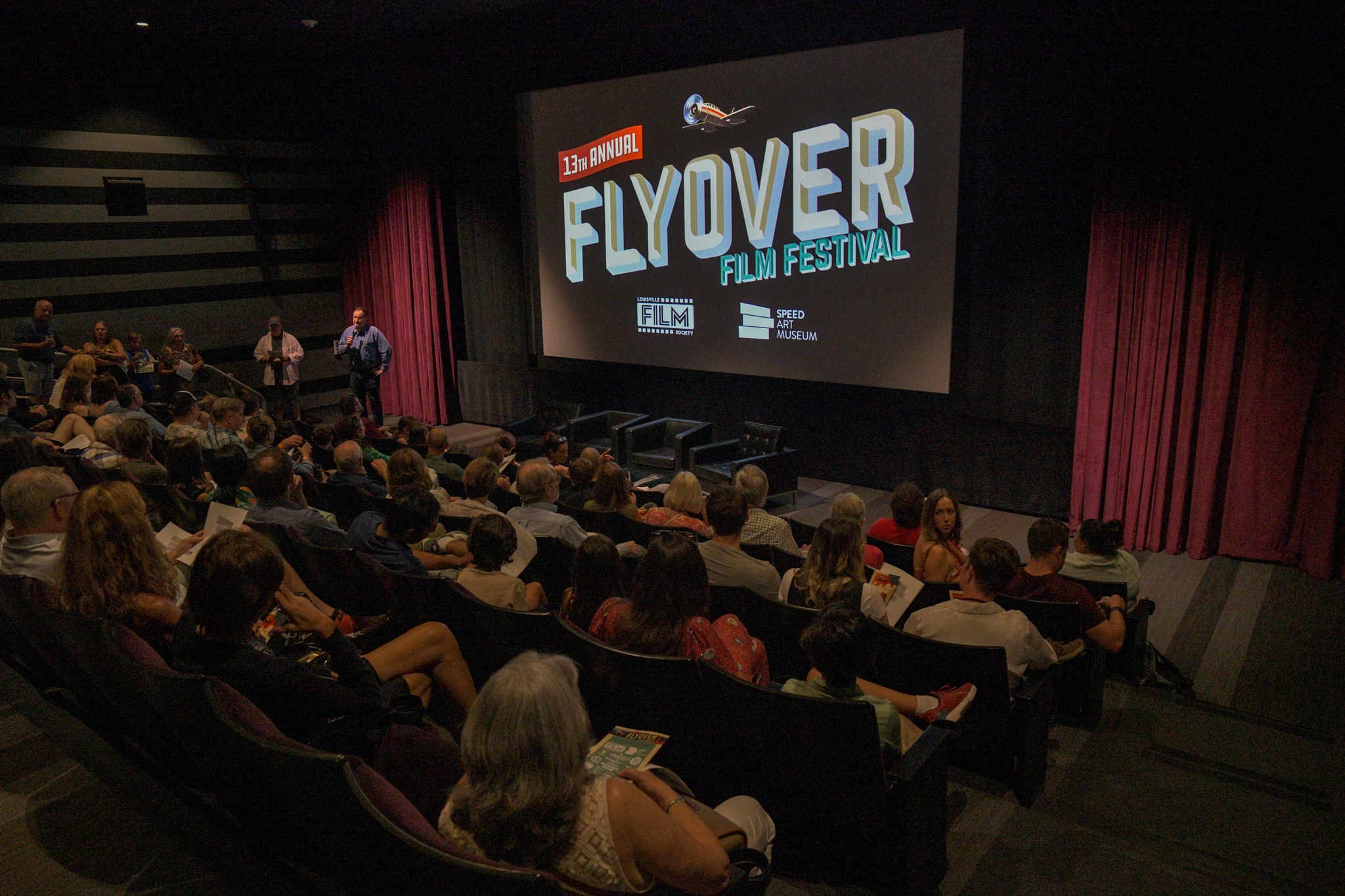 A theater full of seated people is watching a large screen displaying the "13th Annual Flyover Film Festival," celebrating cinema with logos for the Louisville Film Society, Speed Art Museum, and other sponsors.