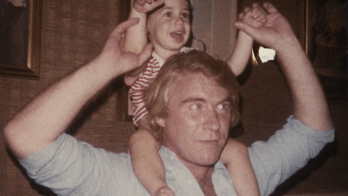 A man with blond hair carries a smiling child on his shoulders, the child donning a red and white striped shirt. 