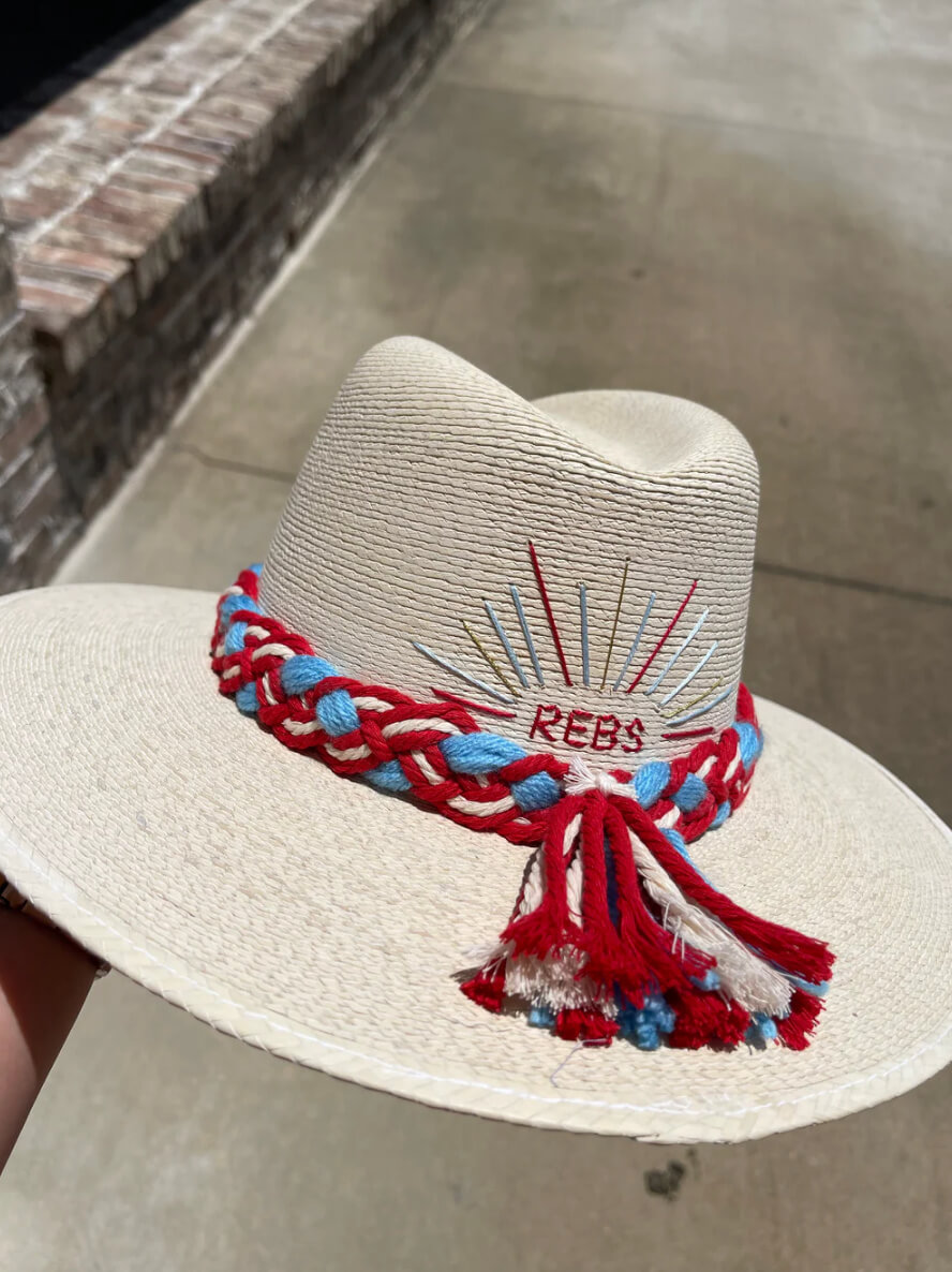 A white hat with colorful braided trim in red, blue, and white, featuring an embroidered design with the text "REBS." The background is a concrete path with a partial brick wall visible.