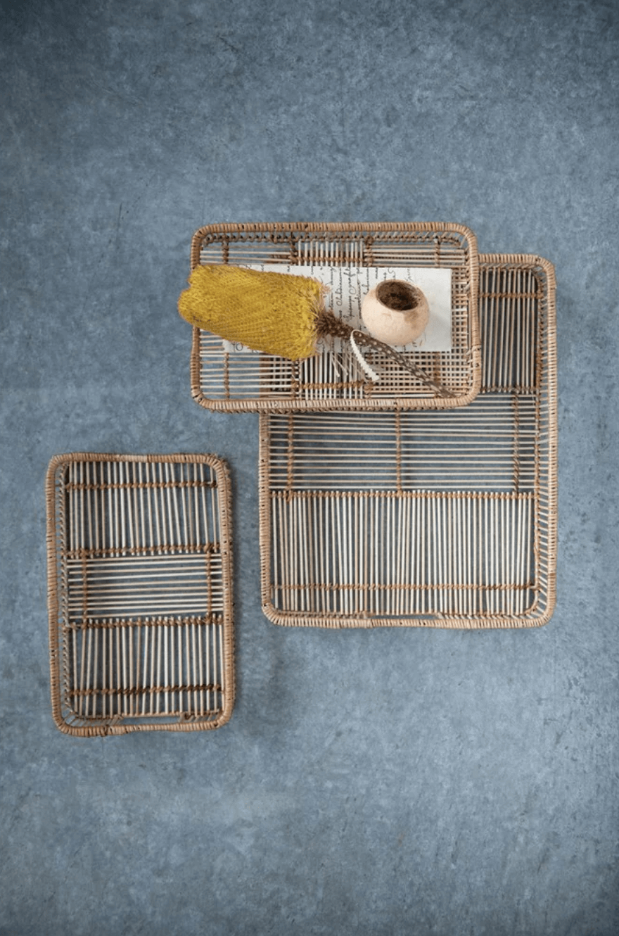 Three rectangular wicker trays of varying sizes are arranged on a gray surface. The largest tray holds a yellow natural sponge, a small bowl, and a brush.