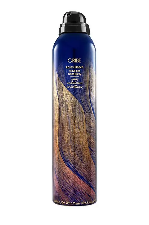 A tall, dark-colored spray bottle labeled "Oribe Après Beach Wave and Shine Spray," featuring a design resembling flowing hair. 