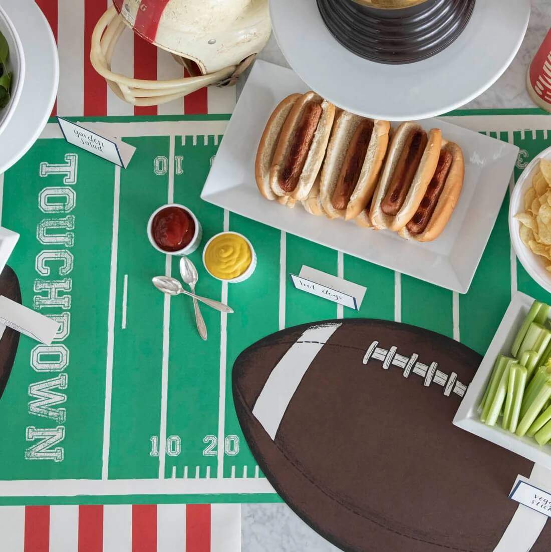 A table with a football-themed tablecloth is set with hot dogs, chips, celery sticks, condiments, and a football cutout. A football helmet completes the festive setup.