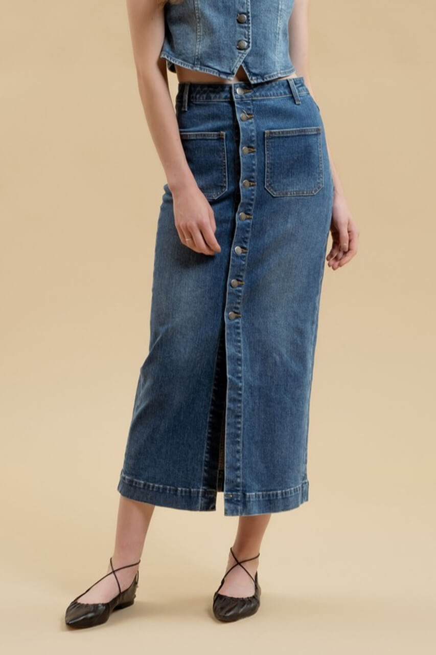 A person wearing a button-up denim midi skirt and a matching denim top stands against a beige background. The outfit, perfect for September in Nashville, is complemented by black flat shoes with ankle ties.