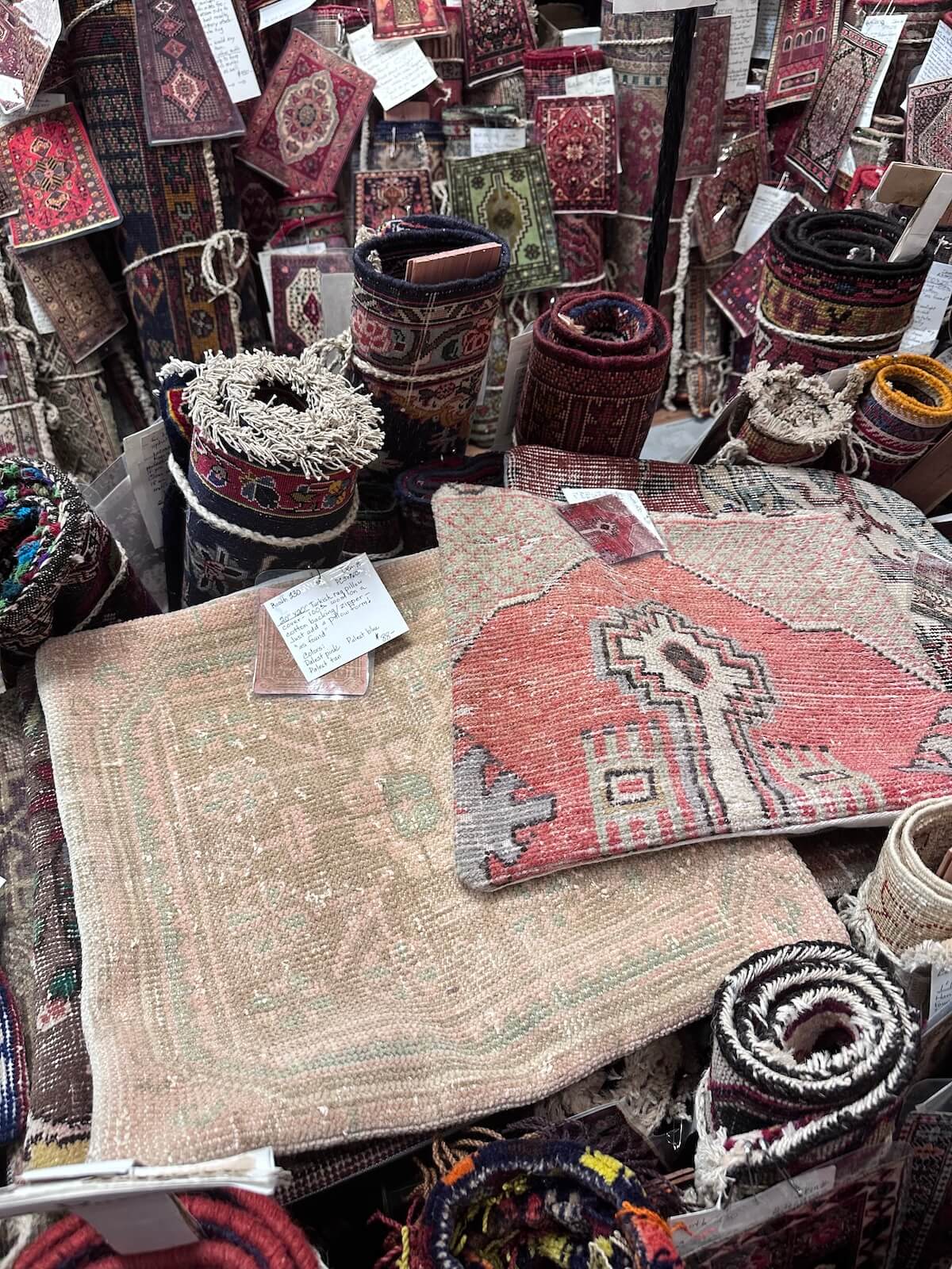 Various rolled and folded patterned rugs and tapestries are displayed with tags attached to some pieces.