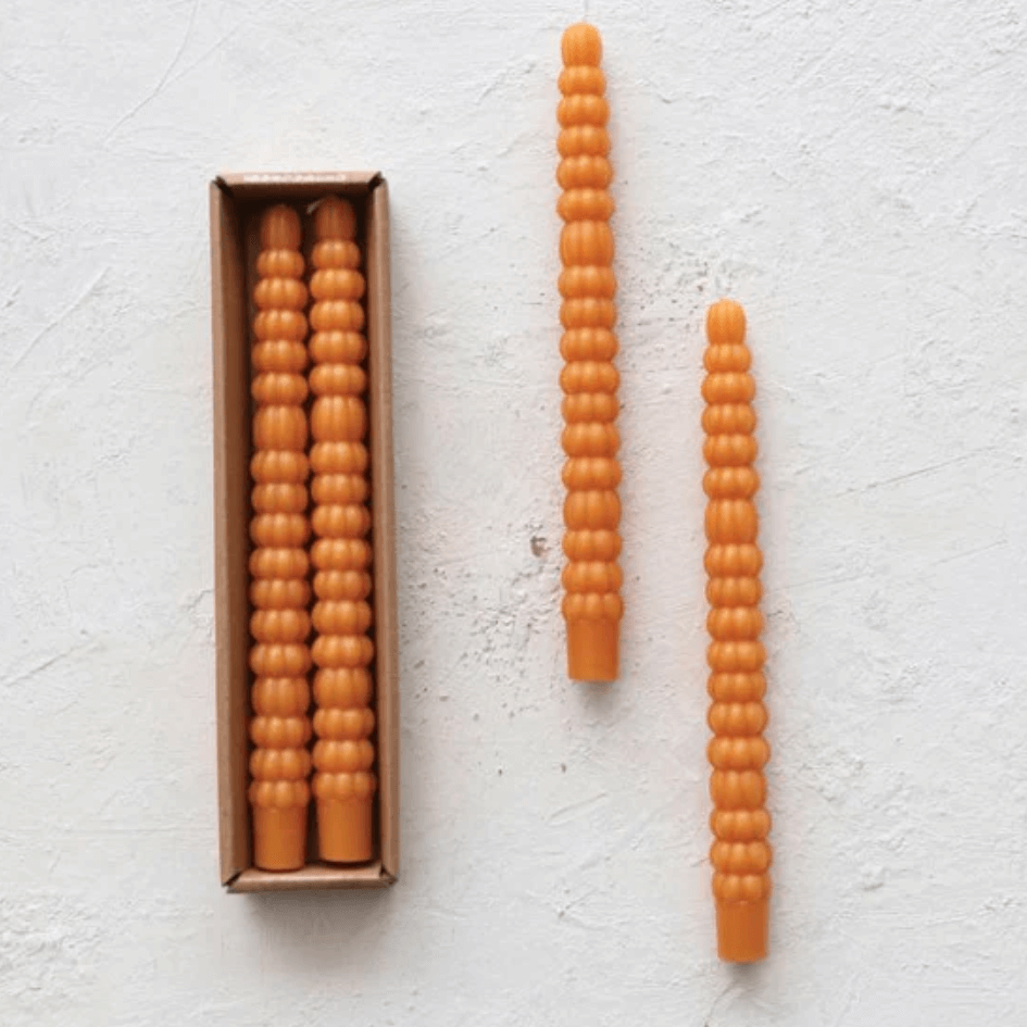 Two orange, ribbed candles with a matching box rest elegantly on a textured white surface, reminiscent of cozy October evenings in Nashville.