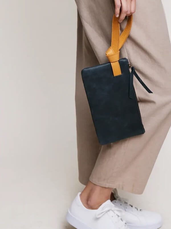 A person stylishly holds a black clutch with a yellow handle, while wearing beige pants and white sneakers.