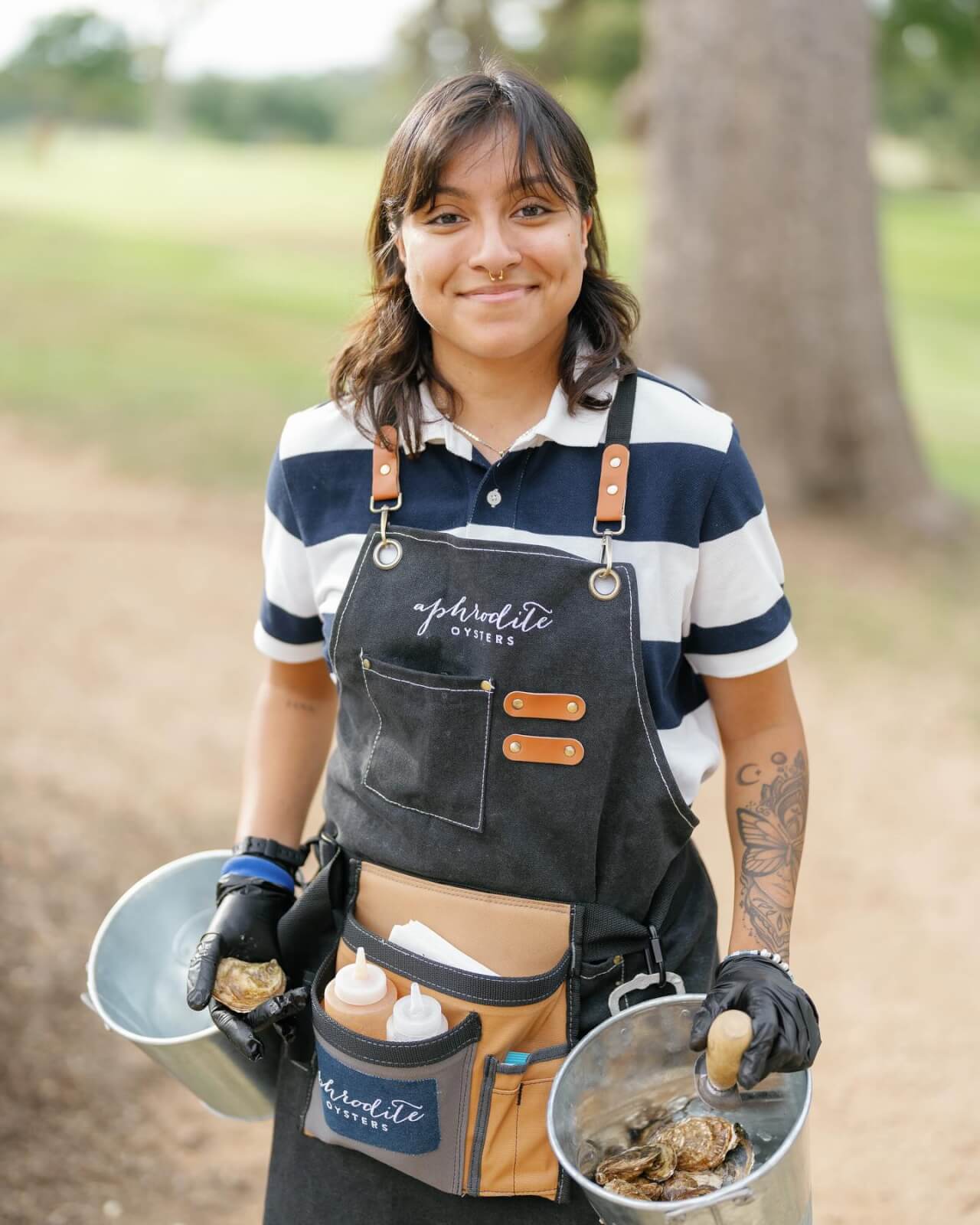Person wearing a striped shirt and apron, holding a metal bucket with oysters. They have tattoos on their arm, a utility belt with condiments, and are standing outdoors.