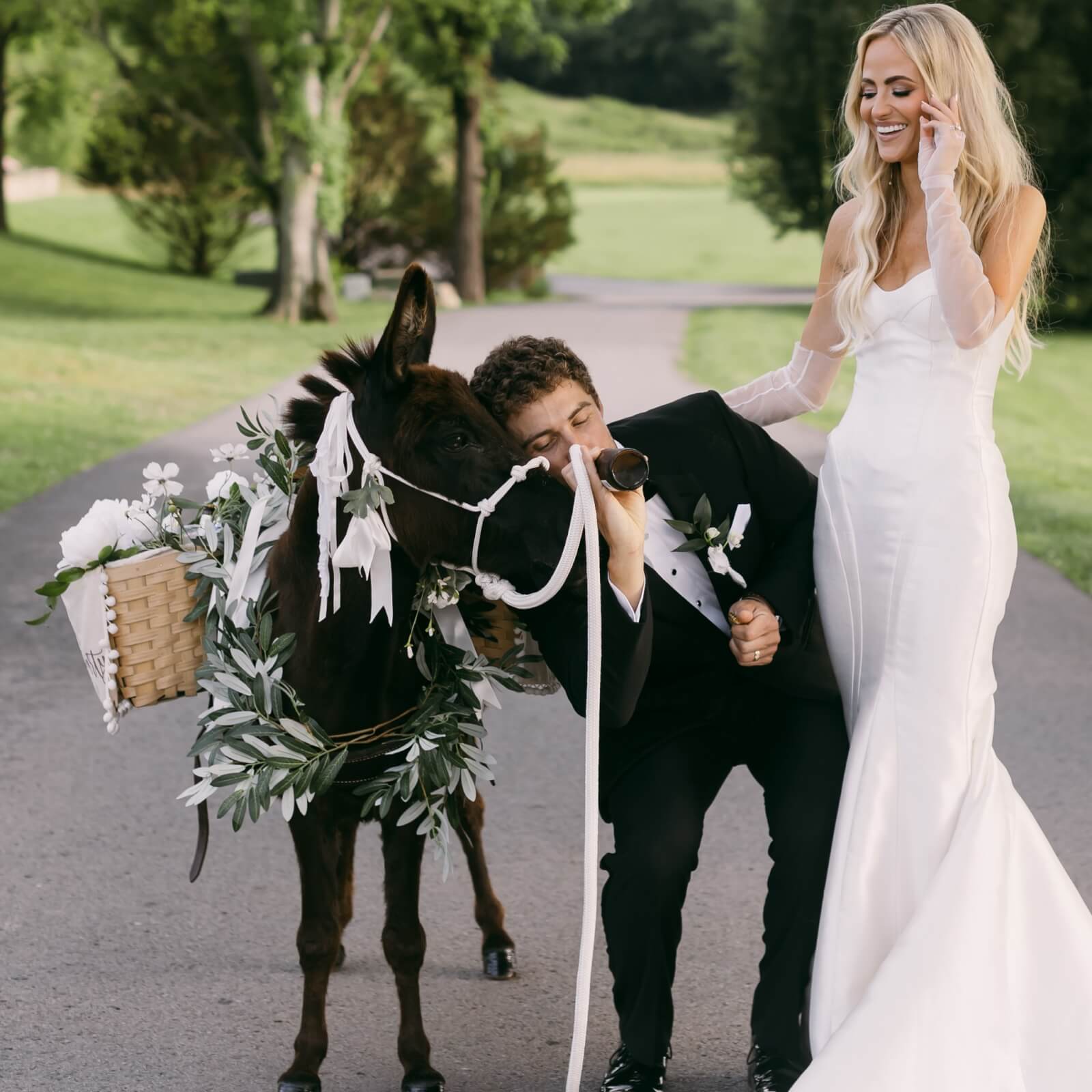 Bride and groom stand on a path beside a ribbon-adorned donkey, with the groom holding a drink and the bride smiling while touching her hair. 