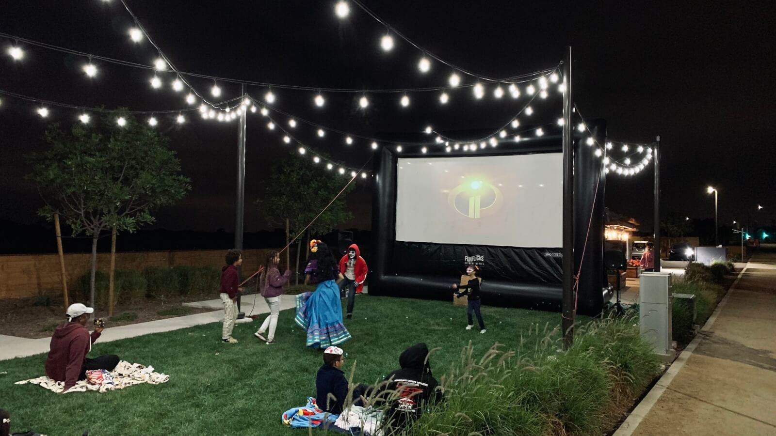 Experience the magic of an outdoor movie night with Nashville Party Rentals. People gather under string lights, watching a film on a large inflatable screen. Some individuals sit on the grass while others stand nearby.