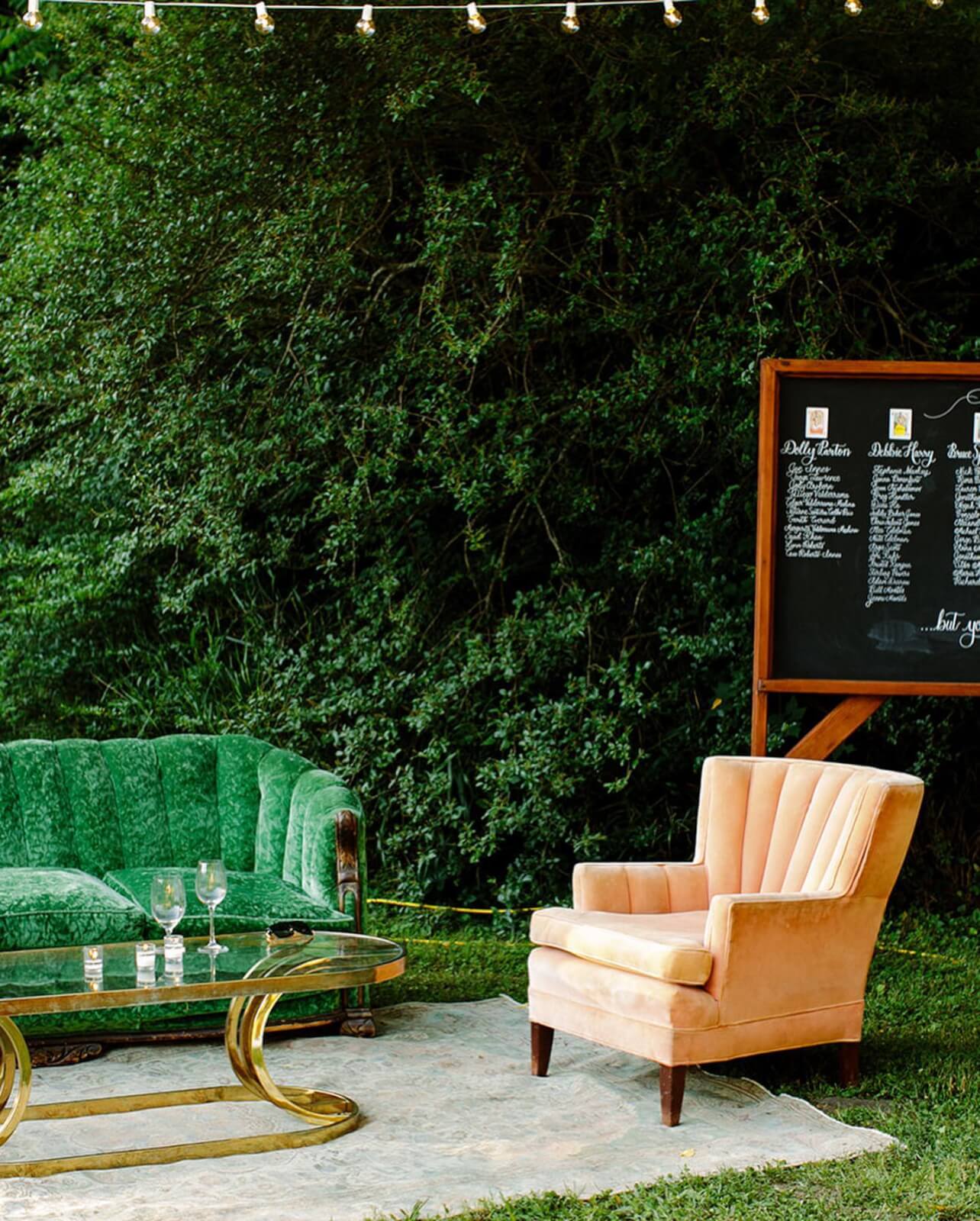 Elevate your next event with our outdoor seating area featuring a green sofa, pink armchair, and glass coffee table. A menu board stands in the background, complemented by string lights overhead.