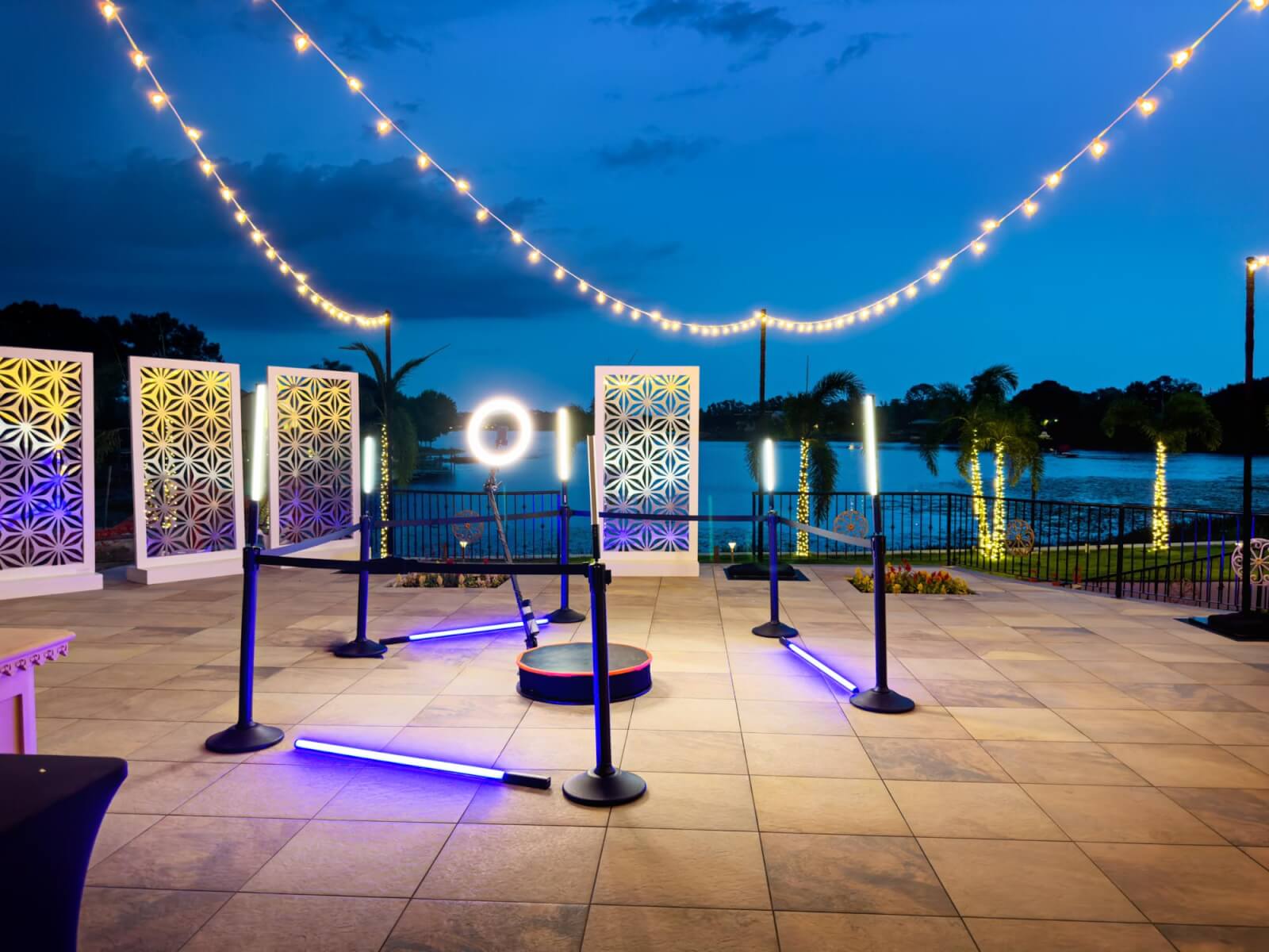 Elevate your event in our outdoor space at dusk, featuring patterned partitions, string lights, neon light decor, and a stunning view of the lake in the background.