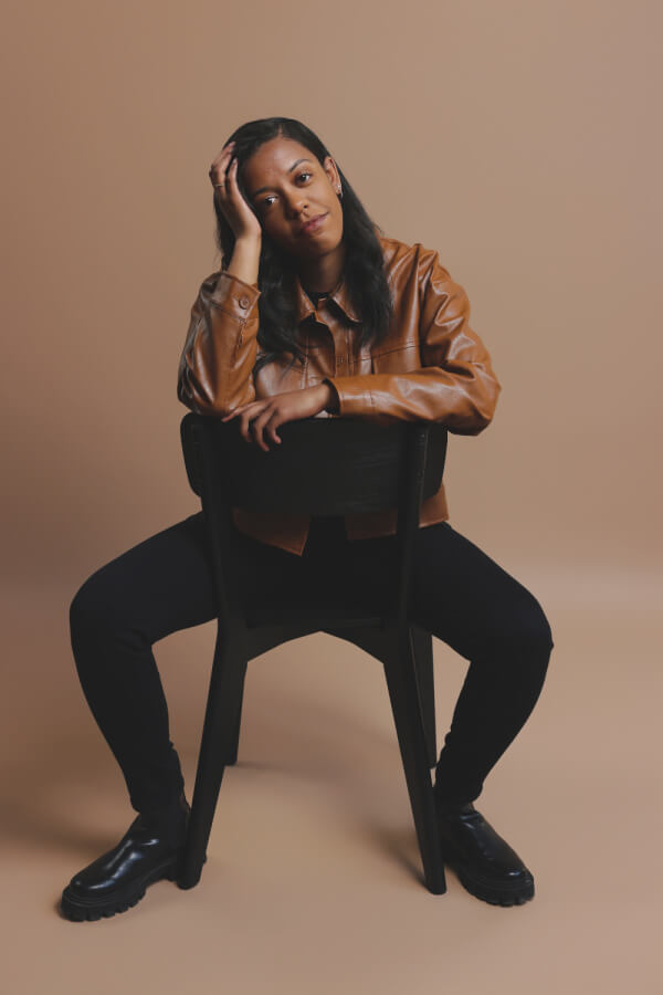 Alex is sitting on a chair backward, resting their head on one hand, wearing a brown jacket, black pants, and black shoes against a neutral background.