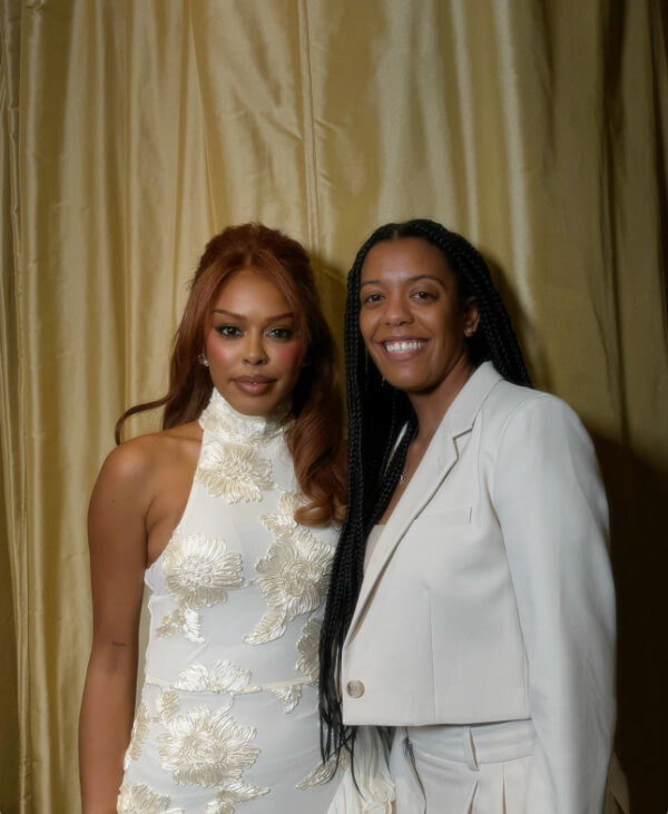 Two individuals stand side by side. Alex Evelyn wears a white halter dress with floral patterns, while the other person sports a white blazer over a top and pants. Their smiling faces light up against the gold curtain backdrop.