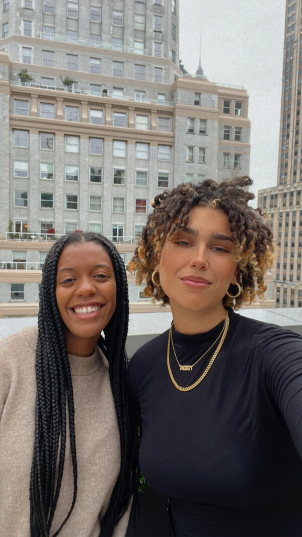 Alex and Evelyn are smiling on a rooftop with the FACE of tall buildings in the background.