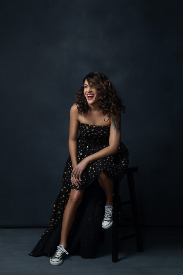 A woman with curly hair in a black dress with yellow stars sits on a stool, smiling and laughing. She wears white sneakers and has a tattoo on her left upper arm. 