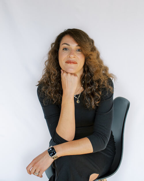 A person with curly brown hair, wearing a black long-sleeve top and watch, sits on a chair with hand on chin, her face thoughtful against a plain white background.