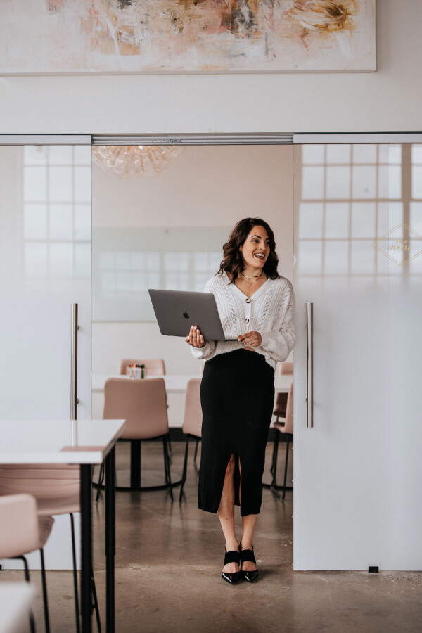 In a modern office, Jackie Gutierrez-Jones stands smiling with confidence as she holds a laptop, her white sweater and black skirt perfectly complementing the professional ambiance.