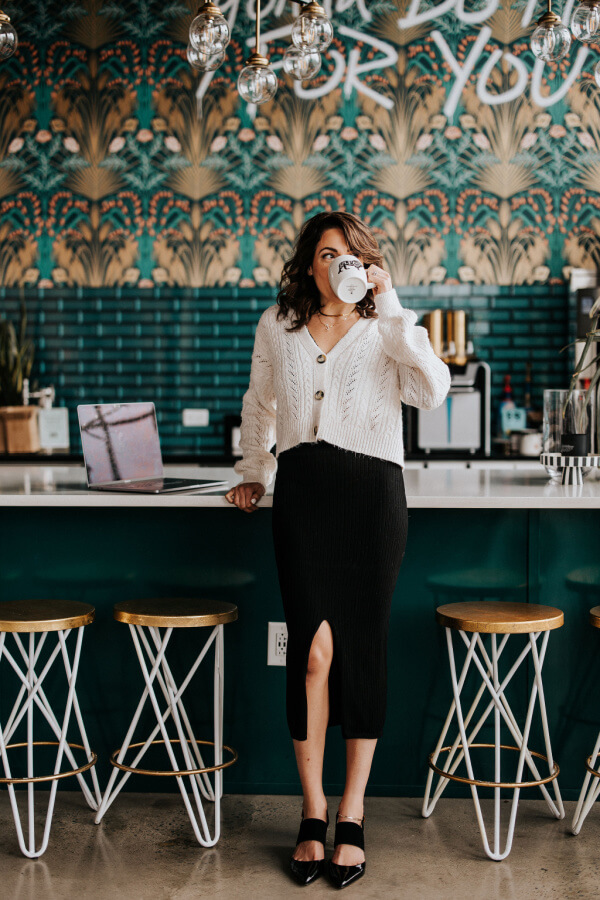 Jackie Gutierrez-Jones, dressed in a white sweater and black skirt, stands at the counter of a stylish café with a teal and patterned wall, drinking from a cup. Her face radiates contentment in the cozy ambiance.