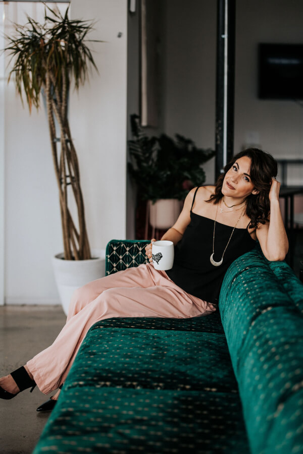 Jackie Gutierrez-Jones, sits on a green patterned sofa holding a white mug. She is wearing a black sleeveless top and light pink pants. A tall potted plant stands gracefully in the background.