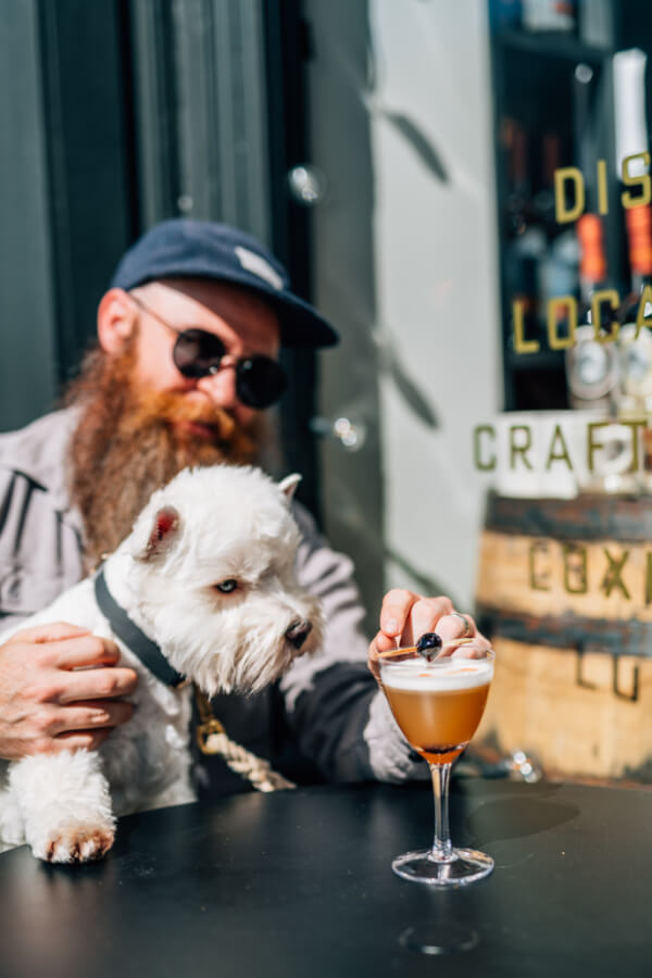 A man with a beard and sunglasses holds a white dog while placing a garnish on a cocktail at an outdoor table, showcasing the charm of the TN Pawsport experience.
