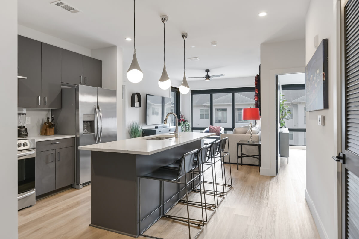 Modern kitchen and living space at 303 31st Ave N in Nashville, TN, featuring an island with bar stools, stainless steel appliances, hanging pendant lights, and a bright living room with large windows in the background.