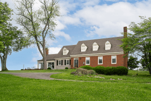 A red-brick and beige-sided house with a steep roof and dormer windows sits on a well-maintained lawn with trees and a curved driveway at 1101 Reed Lane, Simpsonville KY.