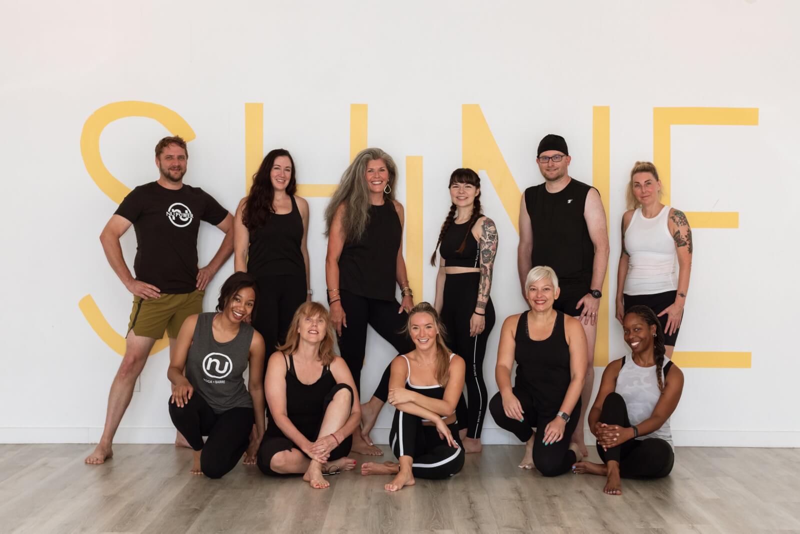 Group of eleven people posing in front of a wall with the word "SHINE" in bold yellow letters, proudly representing the best yoga community.