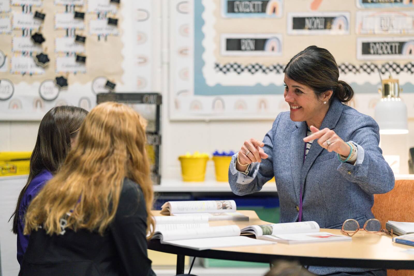 A teacher sits at a desk, smiling and gesturing while talking to two students in a classroom setting at a Nashville private school.