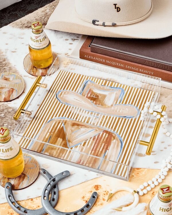 A tray with painted cowboy hats, two beer bottles, coasters, a beige hat, books, horseshoes, beads, and other Western FINDS sits on a cowhide surface.