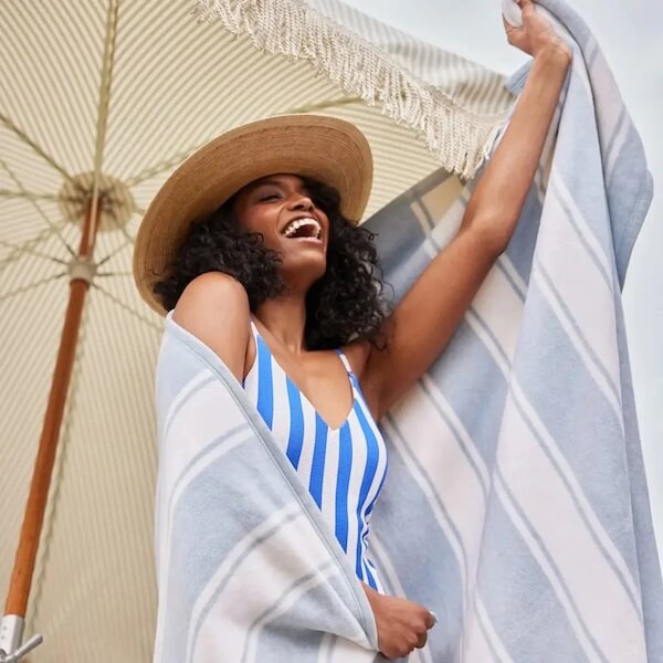 A woman in a blue and white striped swimsuit and straw hat smiles while holding up one of her latest FINDS—a matching striped towel—under a beach umbrella.