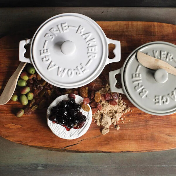 Two ceramic brie bakers with lids—true entertaining FINDS—sit on a wooden board, surrounded by olives, raisins, brown sugar, a round of brie cheese, and a fruit topping.