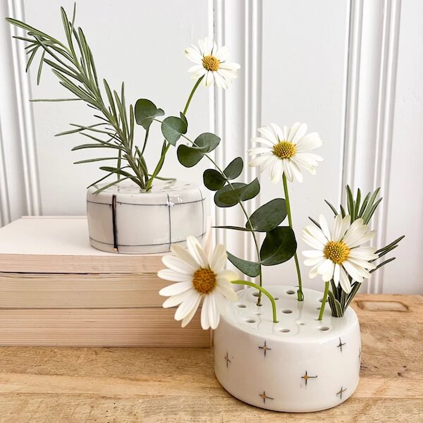 Two small ceramic vases—true FINDS—filled with white daisies, eucalyptus leaves, and rosemary sprigs are displayed on a wooden surface and a stack of books against a white paneled wall.
