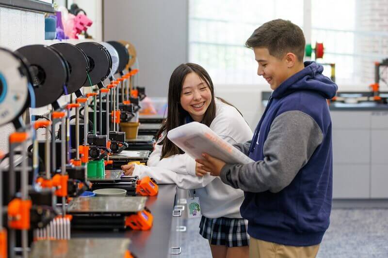 In a Nashville private school, two students interact in a classroom filled with the hum of multiple 3D printers on a long table beside them. One student holds a notebook, and both are smiling.