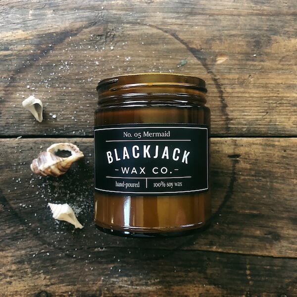 A brown glass jar labeled "Blackjack Wax Co. No. 05 Mermaid" sits on a rustic wooden surface beside two seashells, echoing the charm of seaside FINDS.