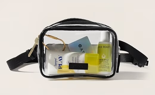 A clear toiletry bag with a black zipper holds various travel-sized skincare and beauty products, including a Supergoop sunscreen, a Juice Beauty serum, and an Ayla card. This sleek clear bag ensures you can easily spot all your essentials at a glance.