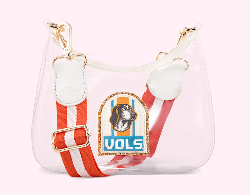 A clear bag with white trim, gold hardware, and a red-and-white striped shoulder strap. It features a paste-over patch with a dog's head and the text "VOLS"