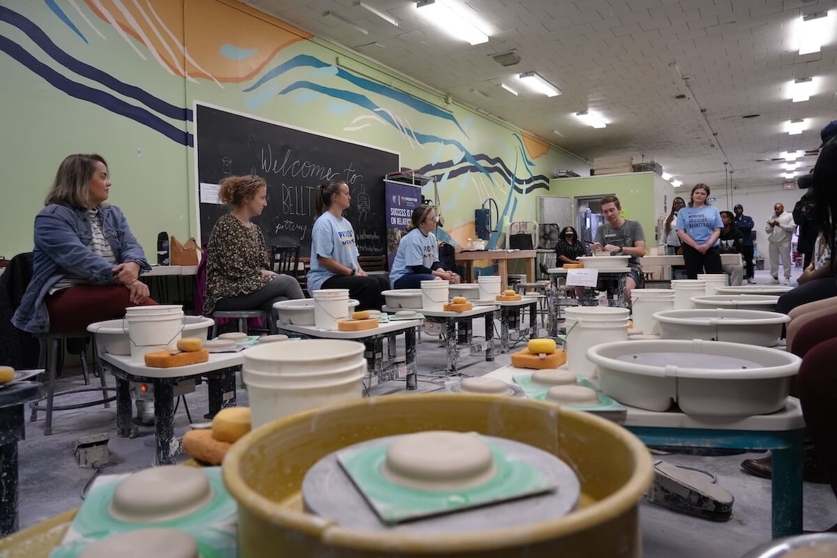 A community of people sit and stand around pottery wheels in a classroom-like setting with a colorful Memphis mural on the wall.