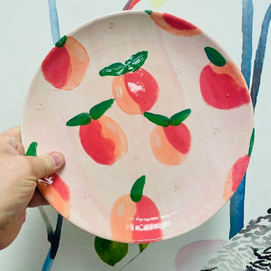 A hand holds a round, ceramic plate from the Memphis Pottery Community, featuring a painted design of multiple red and orange peaches with green leaves on a pale pink background.