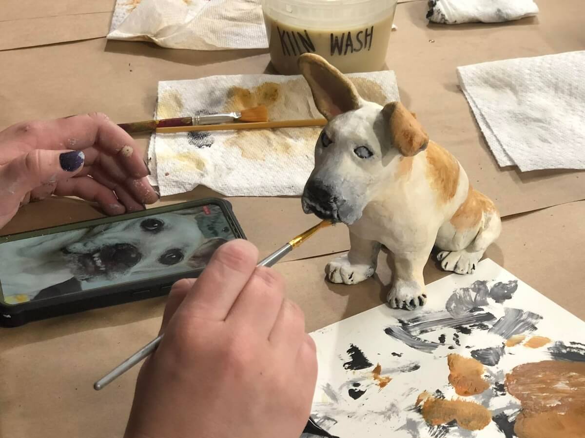 A person paints a dog sculpture on a table, using a photo of the same dog on a smartphone for reference. Various painting supplies and paper towels are scattered around.