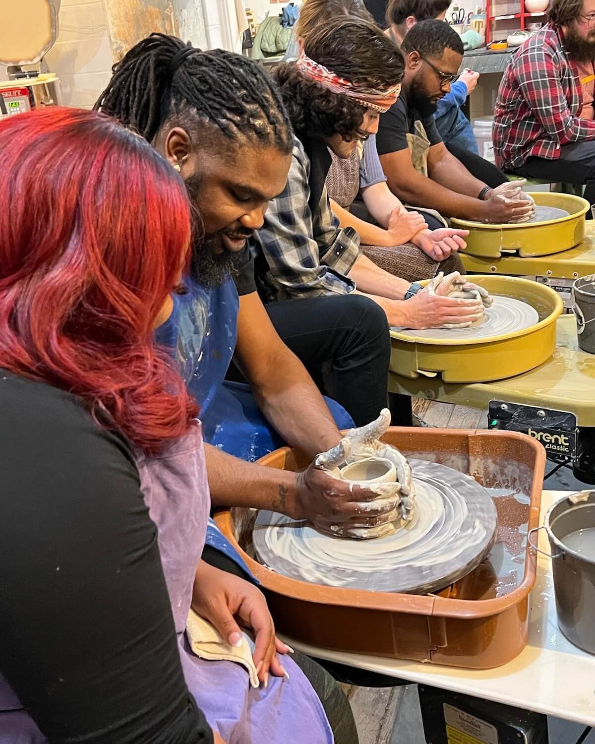 Several people participate in a pottery class, sitting at pottery wheels and shaping clay, with focus on a man and woman in the foreground working together on a piece. 