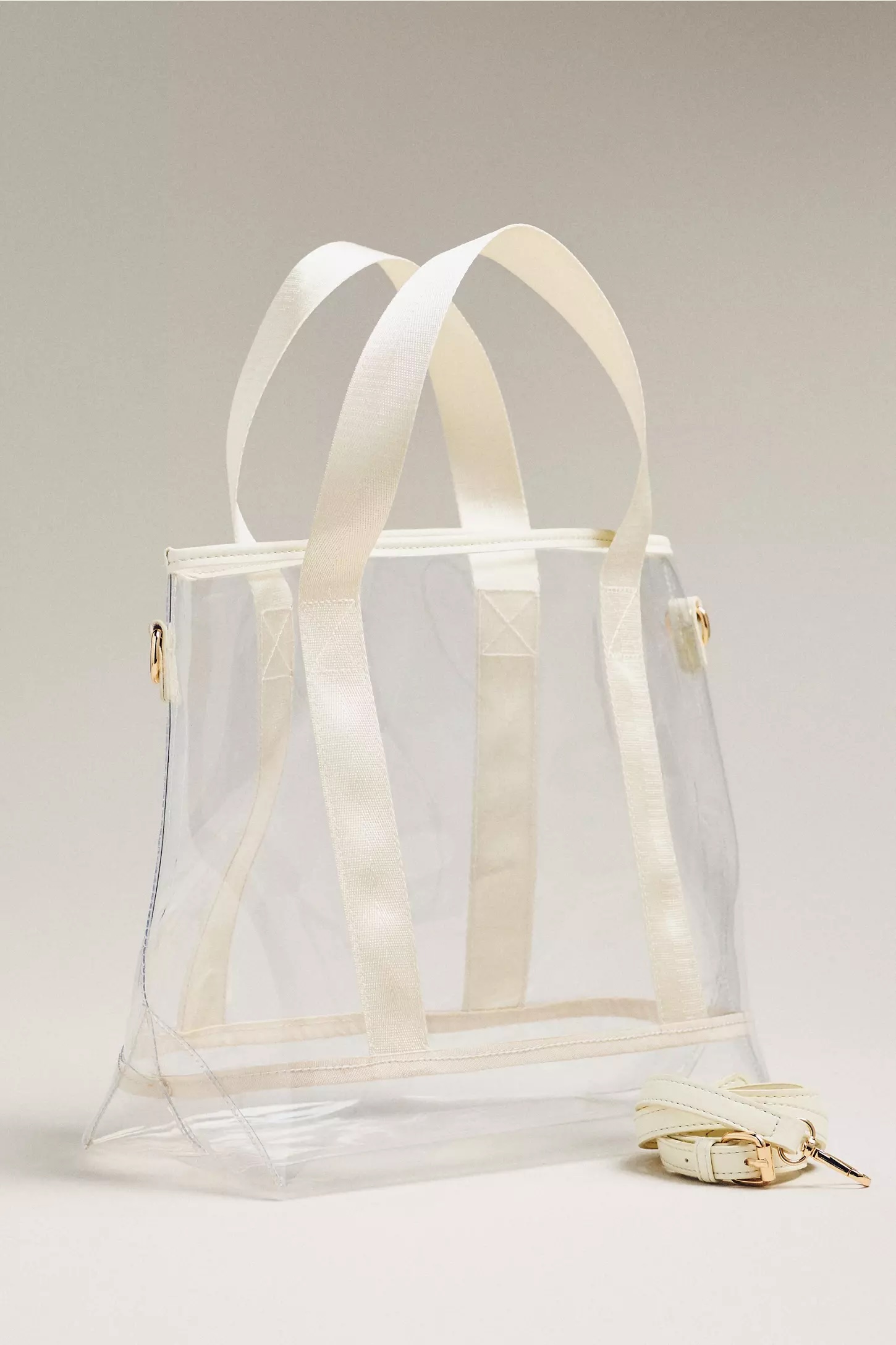 Clear tote bag with white handles and trim, displayed upright on a neutral background, showcases the versatility of clear bags. A matching detachable strap is placed in front for added style and convenience.