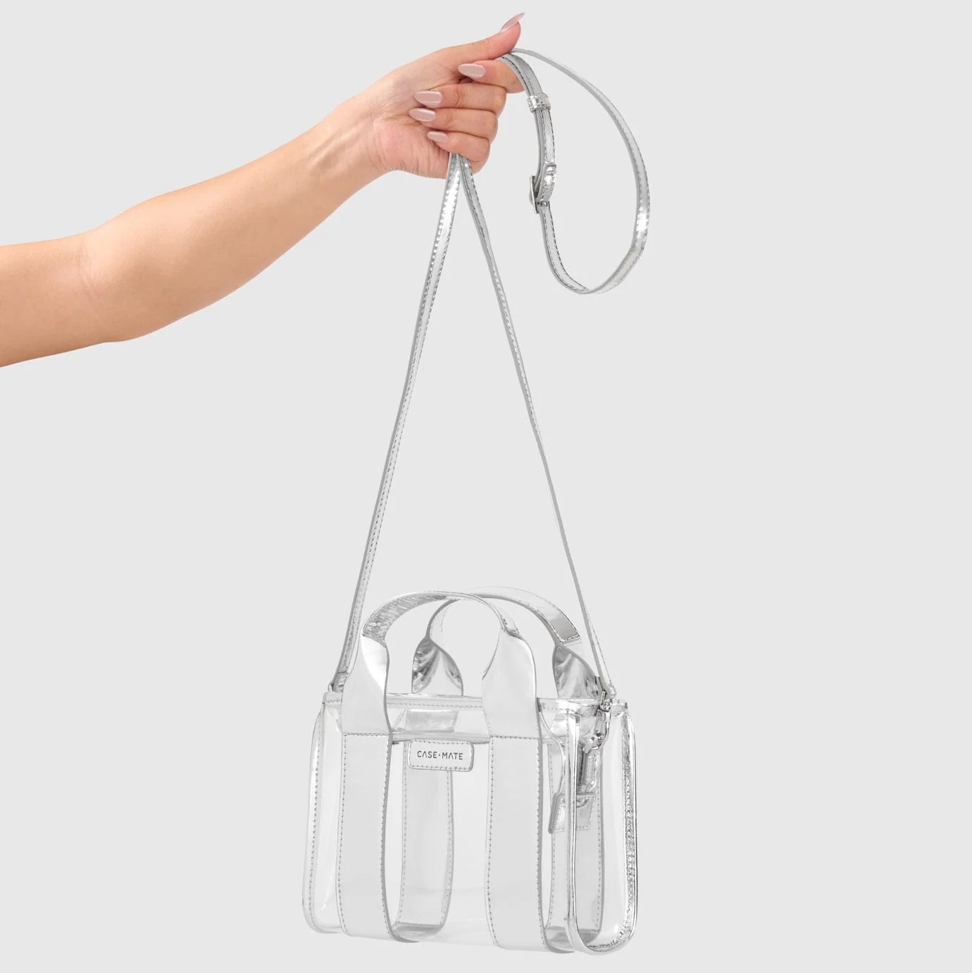 A hand holds a chic, rectangular clear crossbody bag with silver straps and trim against a plain gray background, showcasing the sleek design of clear bags.