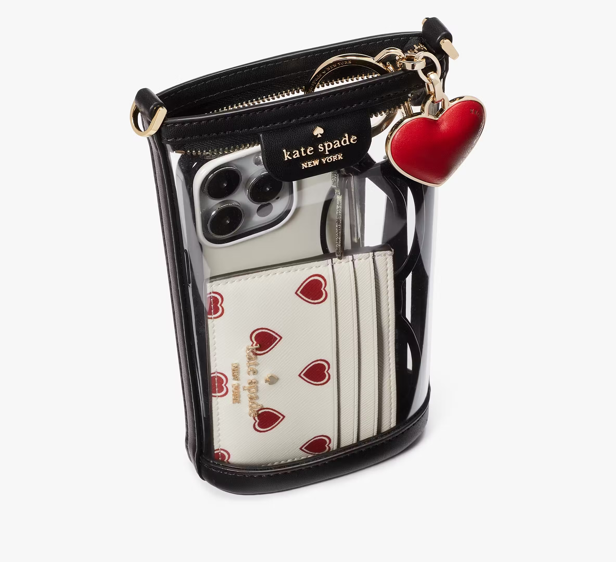 Chic Kate Spade clear bucket bag with black trim, perfect for fans of clear bags; holds an iPhone, white wallet with red hearts, and sunglasses. A playful red heart keychain is attached to the handle.